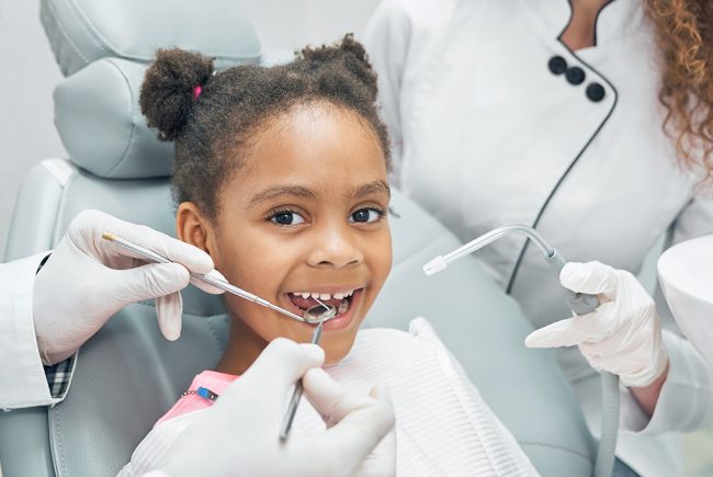 Happy afro american girl sitting in stomatologist chair with open mouth while professional dentist doing regular check up of teeth using dental probe and mirror. Female nurse assisting.