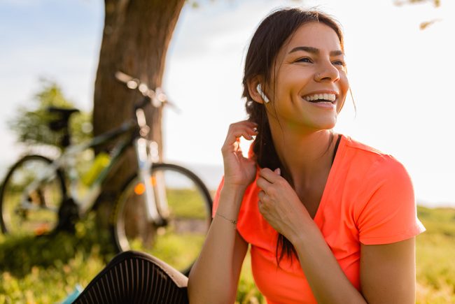 portrait of smiling beautiful woman doing sports in morning in park nature on mat wearing colorful fitness outfit, happy healthy lifestyle, music in earphones, bicycle on background